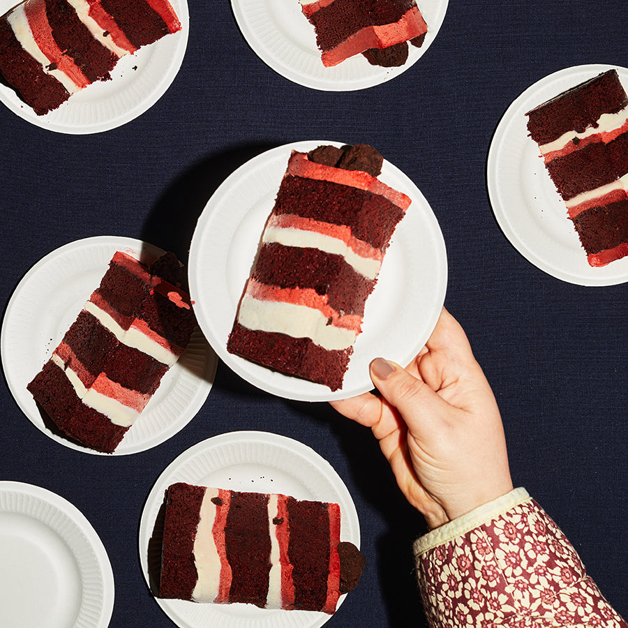 overhead of a dessert table containing several plates of Red Velvet Cake Slices, with a human hand holding up one plate