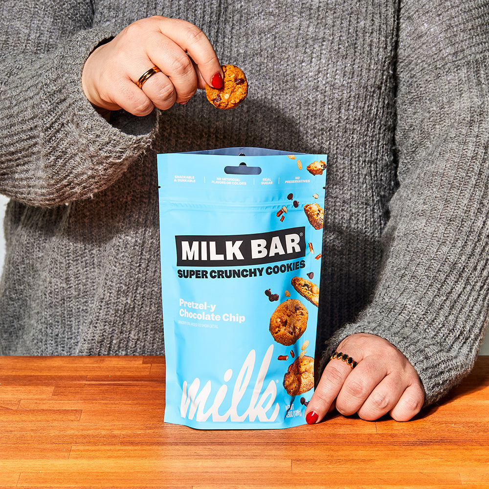 Front view of a bag of pretzel-y chocolate chip crunchies, with a human hand holding up an individual cookie above the bag